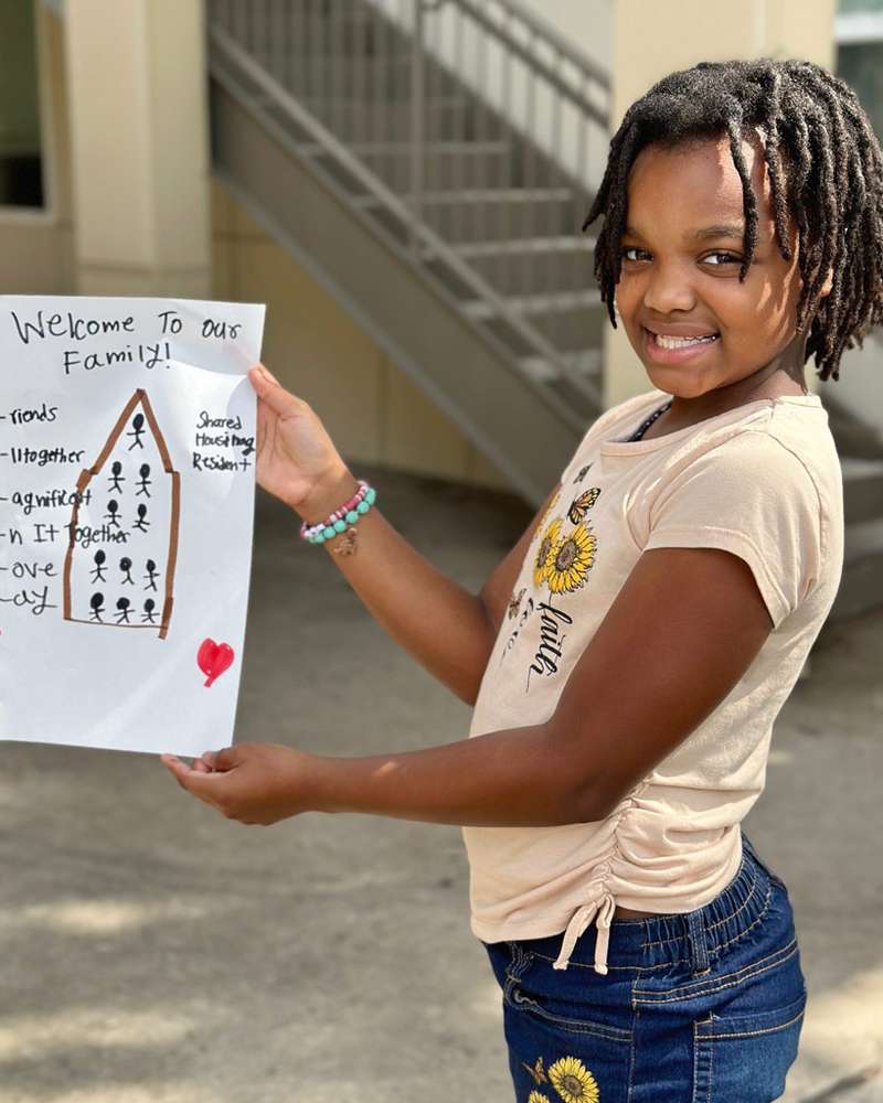 A smiling girl holding up a drawing titled “Welcome to our family!” with a house full of stick figures