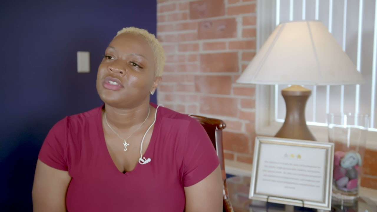A woman in a pink top being interviewed in a room with a lamp
